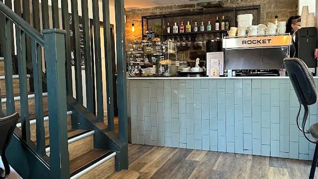 Cozy interior of Joni Grocers with a coffee machine, shelves of goods, and a staircase leading to the upper level.