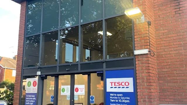 Tesco Superstore entrance with large glass doors, red brick facade, and signage for opening hours and services.