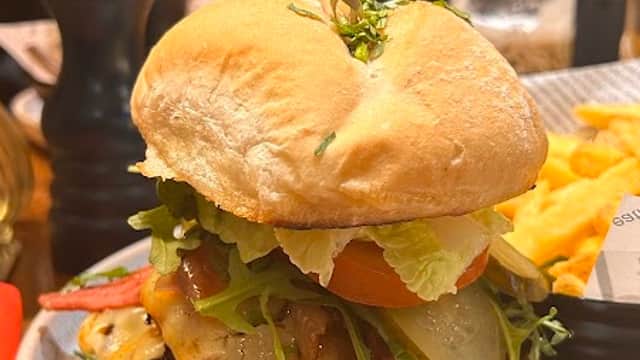 A gourmet burger stacked with bacon, cheese, lettuce, and tomato, served with fries and a flower centerpiece.