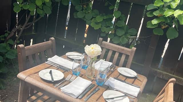 Wooden table set for dining with plates, cutlery, and a flower vase, surrounded by greenery in a cozy outdoor setting.