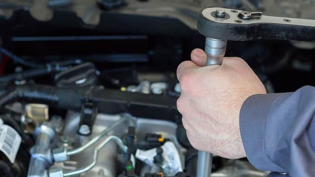 Hands using a ratchet tool on an engine, with various automotive components visible in the background.