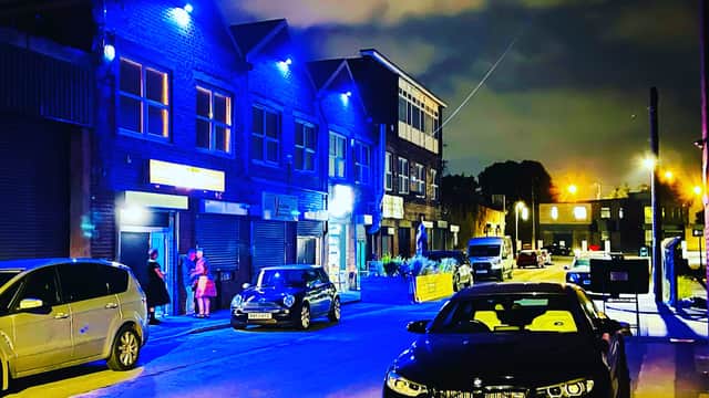 Nighttime scene of Tapestry Arts bar with blue lights, parked cars, and people outside in Dudley Hill, Yorkshire.