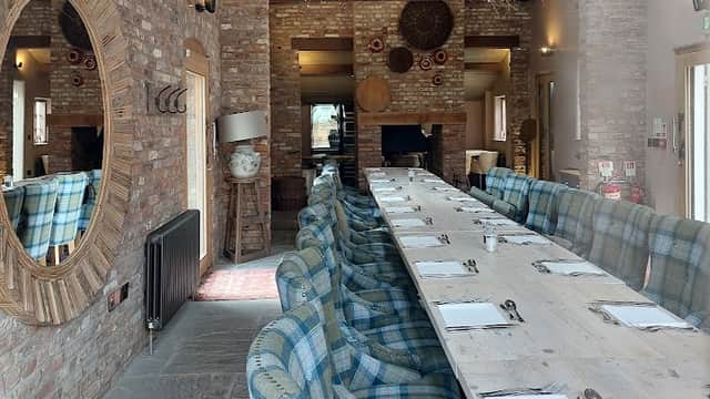 Long wooden dining table with blue plaid seating, exposed brick walls, and decorative lighting in a cozy restaurant interior.