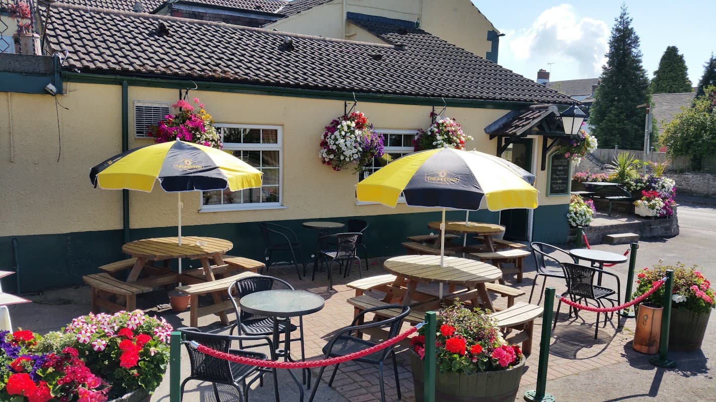Yellow and black umbrellas shade wooden tables outside the Cross Keys Inn, surrounded by vibrant flower displays.