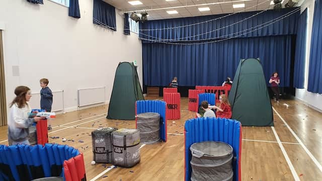 Community center hall with colorful barriers, tents, and children playing in a Nerf gun battle setup.