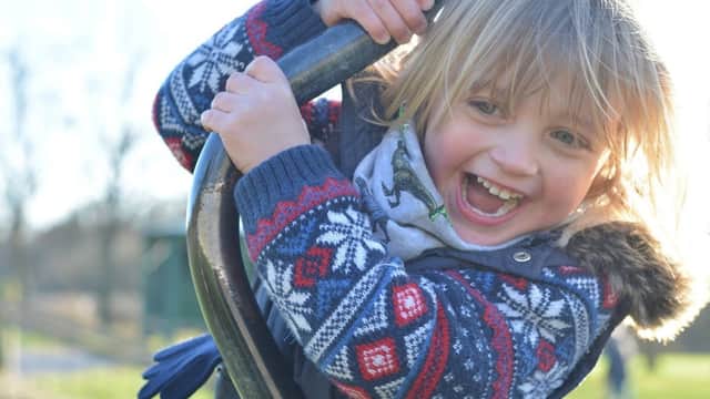 A young child with blonde hair smiles joyfully while climbing on a playground structure, wearing a colorful sweater.