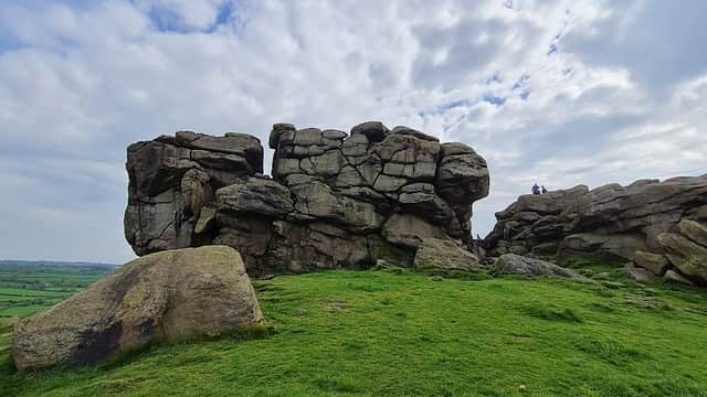 Large rocky outcrop on green grass with cloudy sky, featuring two people near the top of the crag.