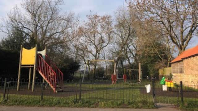 Playground with a slide, swings, and climbing frame surrounded by trees and a low fence in Tockwith, Yorkshire.