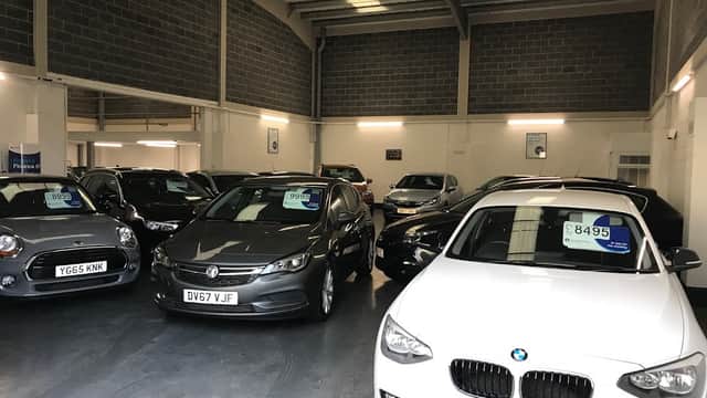 Car dealership interior with various models on display, price tags visible on several vehicles.