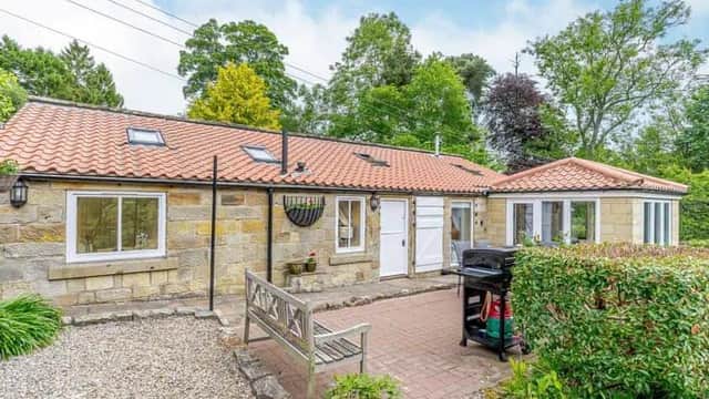 Charming stone cottage with a red-tiled roof, surrounded by greenery and a paved patio area with outdoor seating.