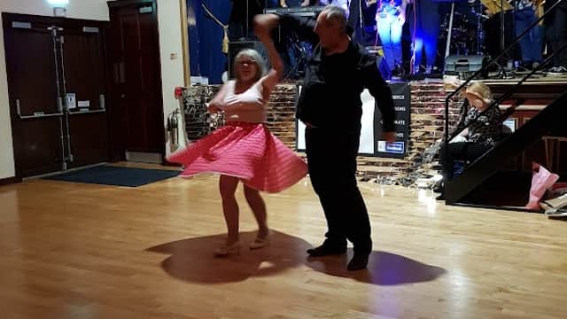 A couple dances on a wooden floor in a hall with a band performing on stage, colorful lights illuminating the scene.
