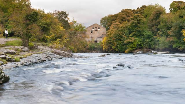 Rushing river flanked by rocky banks and lush trees, with a stone building visible in the background.