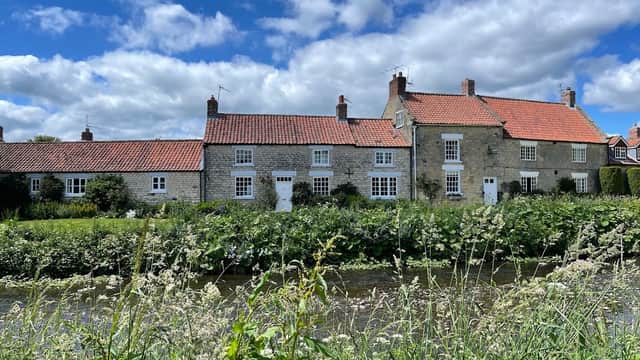 The Fox & Hounds Country Inn features stone buildings with red roofs, surrounded by greenery and a river under a blue sky.