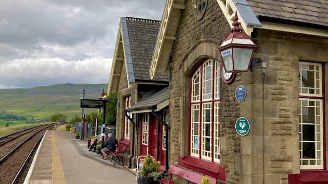 Ribblehead Station features a stone building with red accents, benches, and scenic views of hills under a cloudy sky.