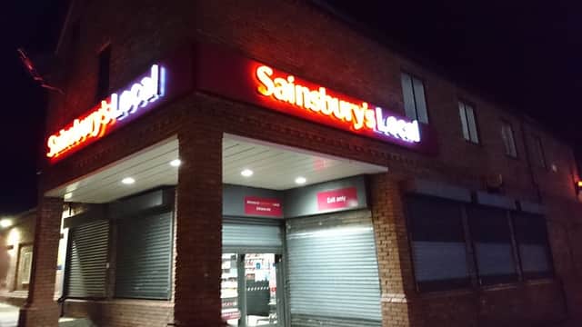 Sainsbury's Local storefront at night, featuring illuminated signage and closed shutters on windows.