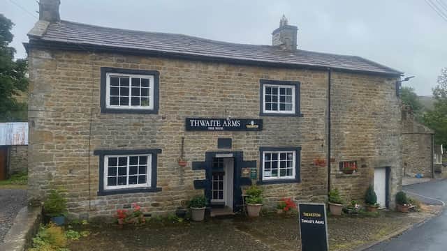 Stone building with "Thwaite Arms" sign, flower pots, and a blackboard menu, set in a rural Yorkshire landscape.