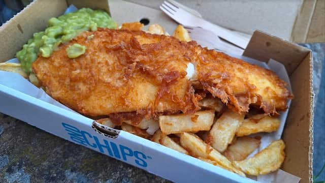 Crispy battered fish served with golden chips and a side of mushy peas in a takeout box.