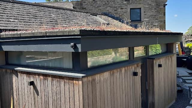 Modern wooden structure with a green roof, surrounded by stone buildings and a landscaped garden in East Marton, Yorkshire.