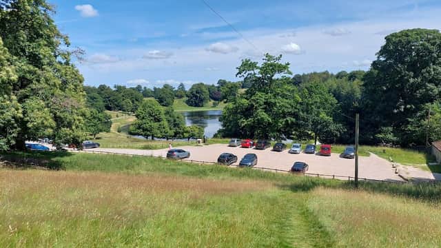 Free parking area with cars, surrounded by lush greenery and a pond under a blue sky with scattered clouds.
