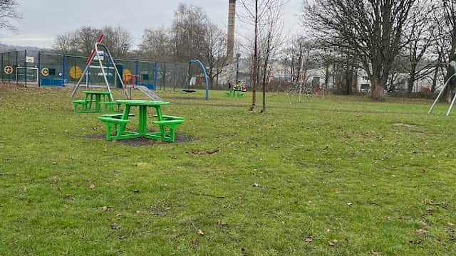 Green picnic tables on grass with playground equipment and trees in Darton Park, Mapplewell, Yorkshire.