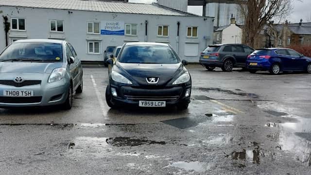 Two parked cars in a gravel lot with puddles, surrounded by buildings under a cloudy sky in Brighouse, Yorkshire.