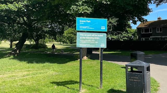 Sign for Graves Park with parking information, surrounded by green grass and trees, near a pathway and a trash bin.