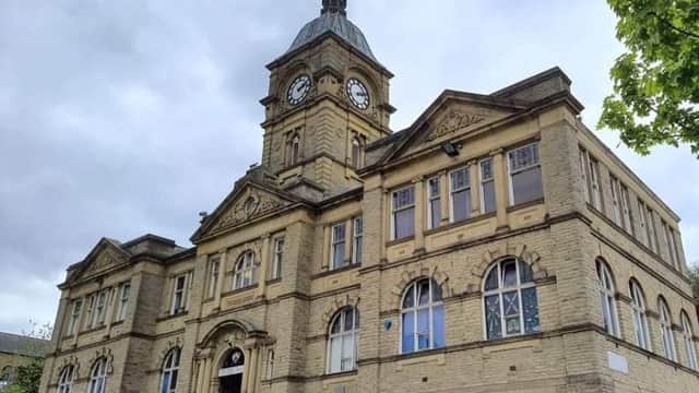 Historic stone building with a clock tower and large arched windows, surrounded by greenery and a cloudy sky.