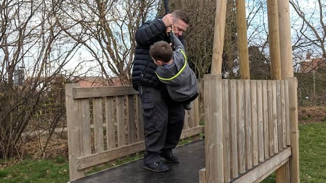 A man helps a child on a wooden play structure with a zip line, surrounded by trees and grassy areas.