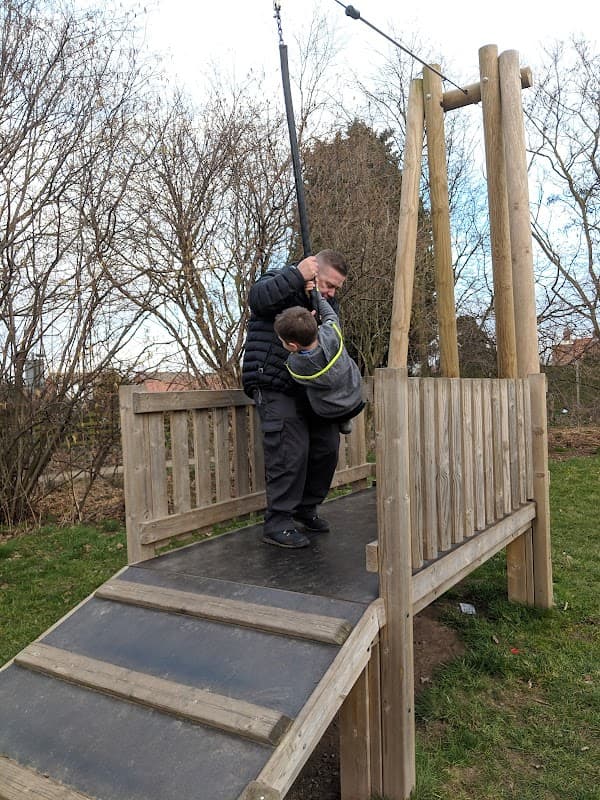 A man helps a child on a wooden play structure with a zip line, surrounded by trees and grassy areas.