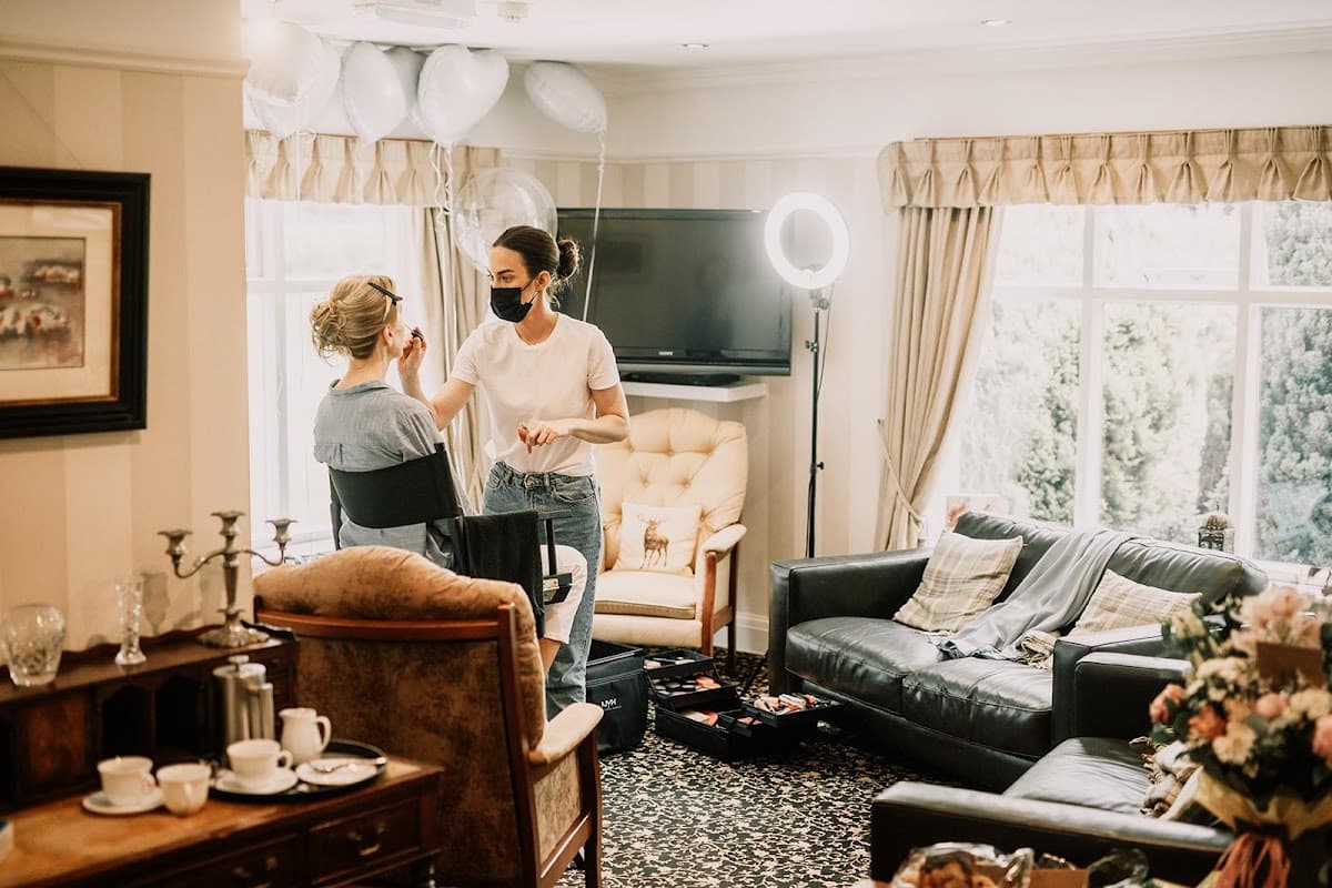 A salon interior with a stylist applying makeup to a client, surrounded by elegant furniture and balloons.