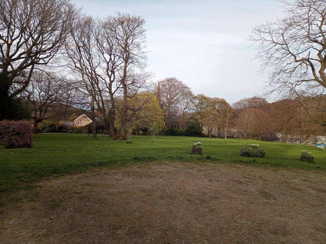 Lush green park with bare trees, a few bushes, and a distant house under a cloudy sky in Meltham, Yorkshire.
