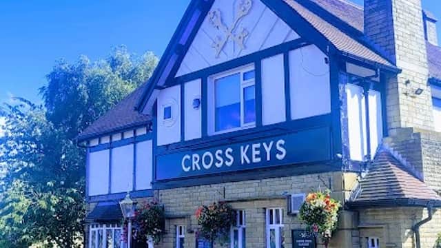 Traditional pub with a sign reading "CROSS KEYS," surrounded by greenery and parked cars under a clear blue sky.