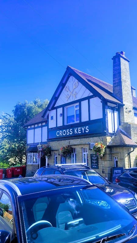 Traditional pub with a sign reading "CROSS KEYS," surrounded by greenery and parked cars under a clear blue sky.