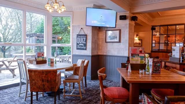 Cozy bar interior with wooden tables, chairs, a TV, and a quiz sign, overlooking a garden area through large windows.