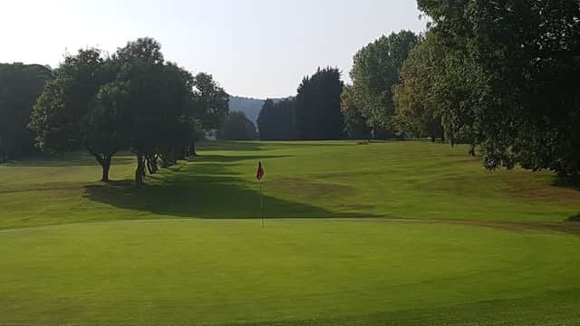 Lush green golf course with a flag on the putting green, surrounded by trees and rolling hills in the background.