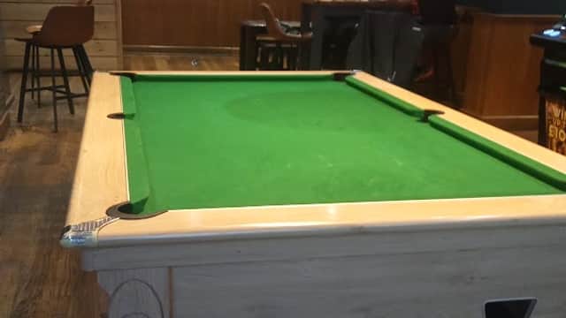 Pool table in a cozy pub setting with a football match on TV and patrons seated at a table in the background.