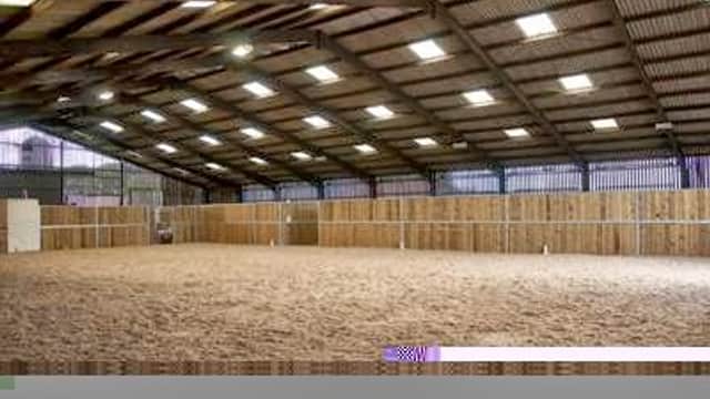 Spacious indoor riding arena with wooden walls and sandy floor, illuminated by overhead lights.