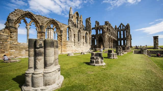 Ruins of Whitby Abbey stand against a dramatic sky, surrounded by grassy fields and a calm water reflection.