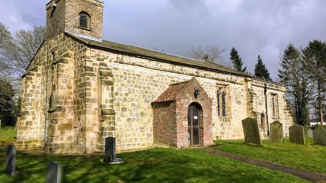 Stone church with a bell tower, arched entrance, and surrounding graveyard in a green landscape under a cloudy sky.