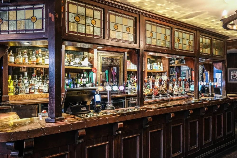 Wooden bar with glass panels, shelves lined with bottles, and taps for various beers at Ye Olde Starre Inne pub.