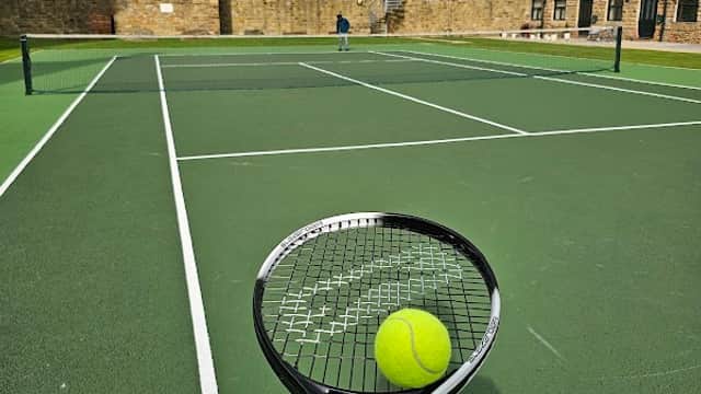 Tennis racket holding a ball, with green courts and historic stone buildings in the background under a cloudy sky.