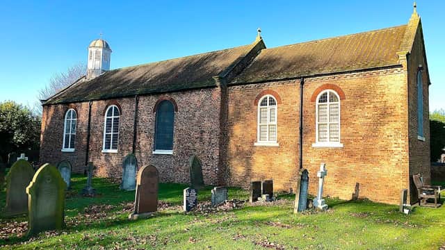 All Saints church in Barlby, Yorkshire, with a brick exterior and gravestones in a grassy cemetery under a clear blue sky.