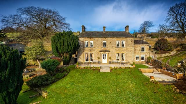 Stone building with large windows, surrounded by greenery and a well-maintained garden in a rural setting.