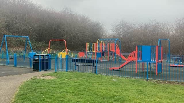 Colorful playground equipment with swings, slides, and climbing structures, surrounded by green grass and trees.