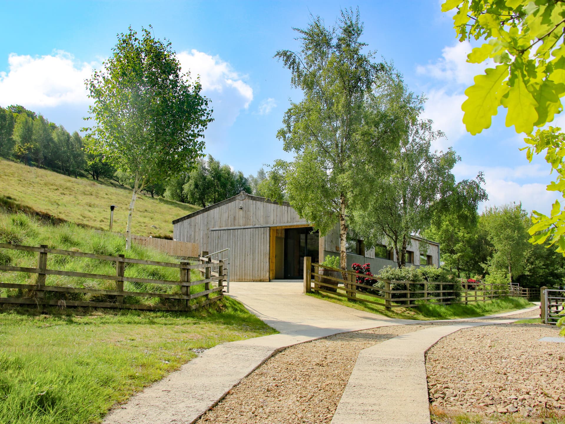 Rustic wooden barn surrounded by lush trees and rolling grassy hills, with a gravel path leading to the entrance.