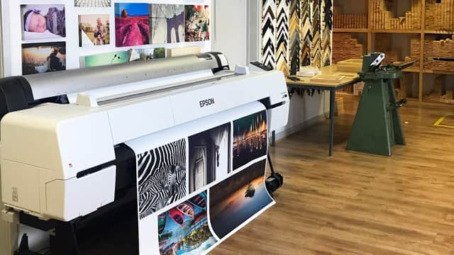 Large printer displaying vibrant images, with wooden shelves and tools in a well-lit workspace.