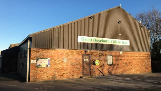 Great Ouseburn Village Hall with a brick facade, metal roof, and signage against a clear blue sky.