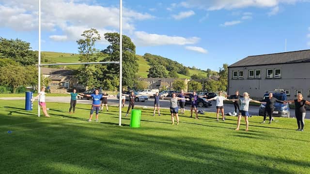 Group of people exercising on a rugby field with goalposts, surrounded by green hills and parked cars.