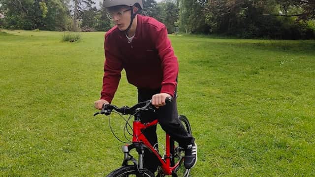 A person in a helmet rides a red bike on grassy terrain, surrounded by trees under a cloudy sky.
