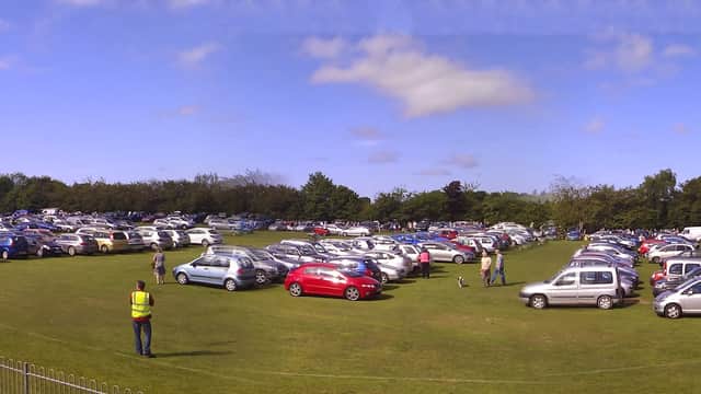 A large grassy field filled with parked cars under a blue sky, with people gathered near picnic tables.